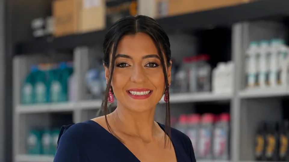 Thaís, sorrindo, no salão da Pactual Auto Peças, com as prateleiras de produtos ao fundo.